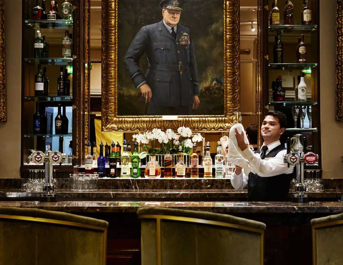 A photo of a smartly dressed bartender making a cocktail behind a grand-looking bar. Above him is a portrait of Sir Winston Churchill.