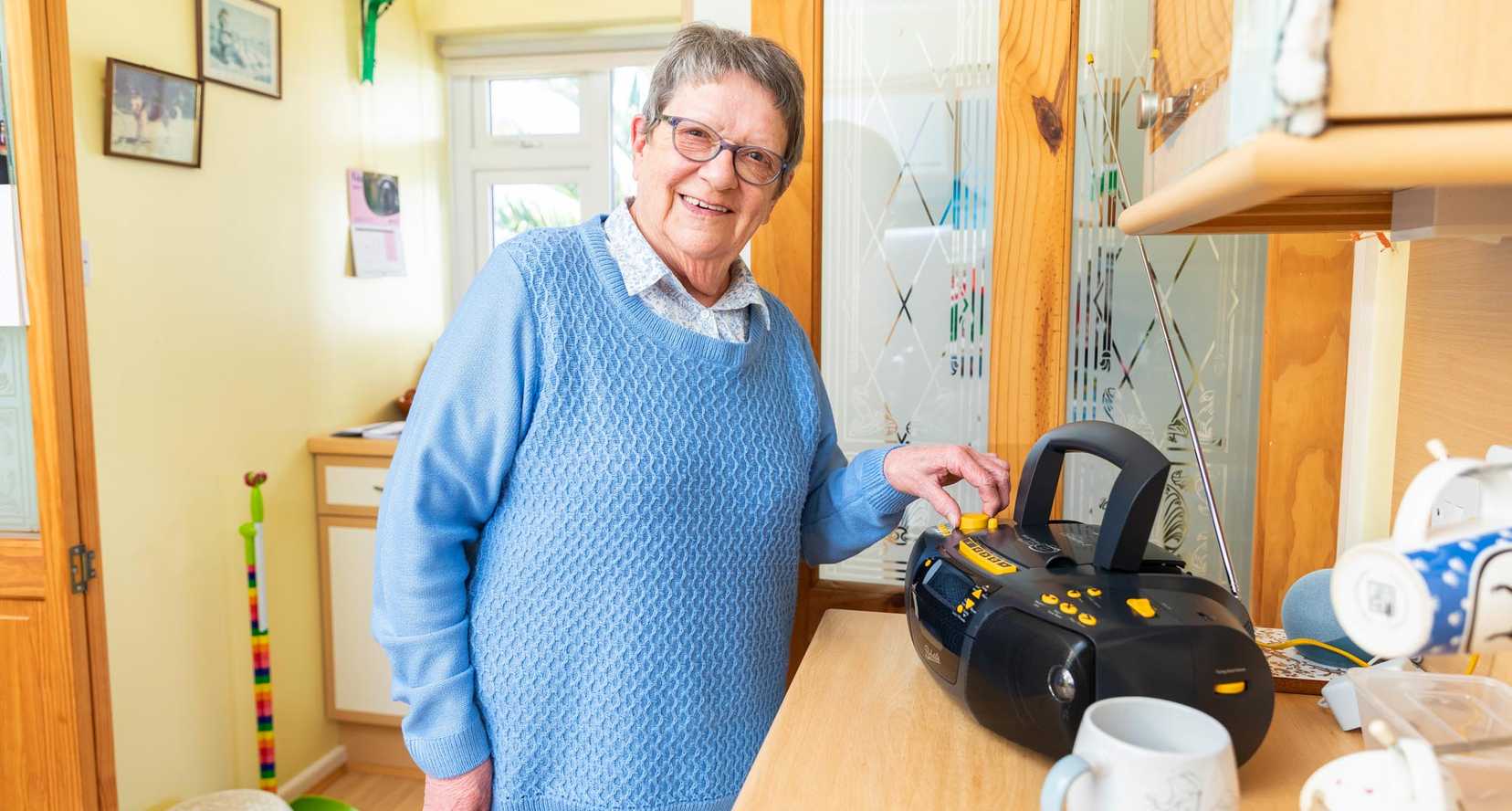 Norma stands in her kitchen smiling as she operates her blck and yellow Concerto 3 radio. She has short grey hair and glasses.
