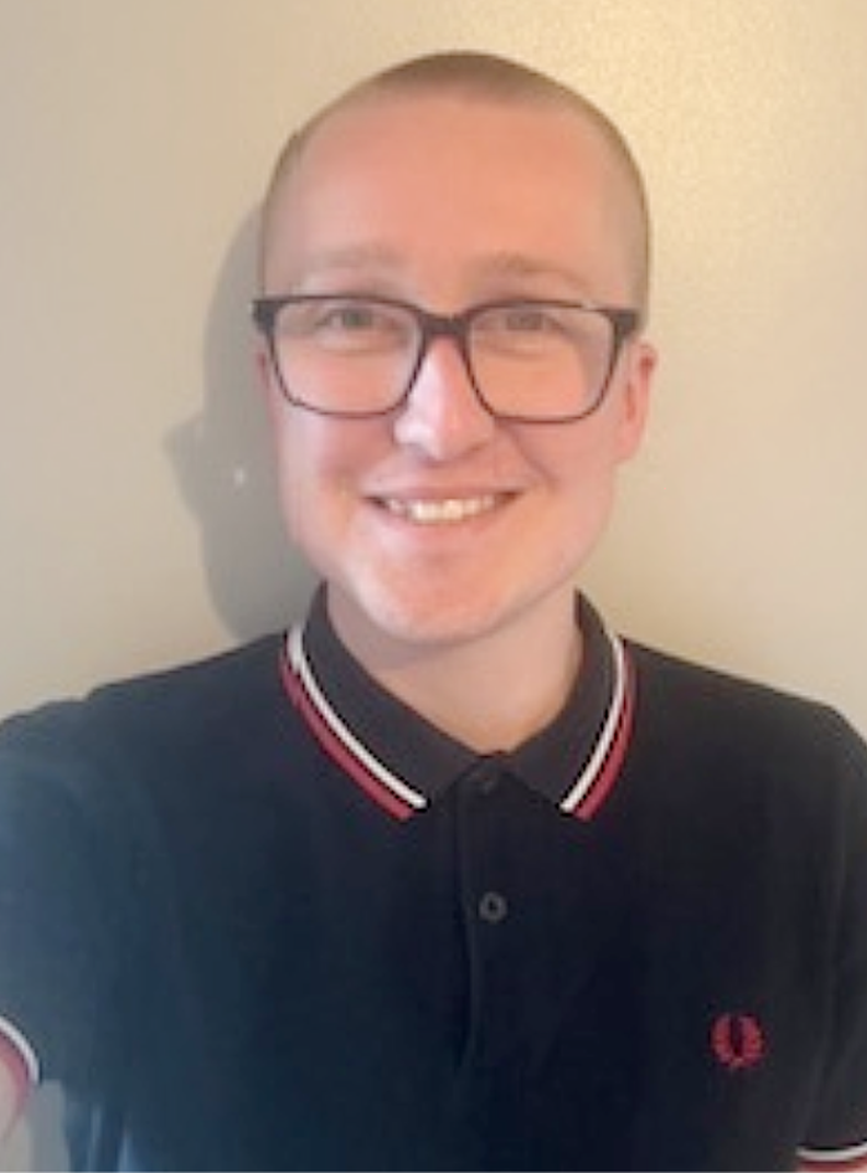 Customer Service Administrator Adam, a young man with a shaved head and glasses, smiling at the camera. He is wearing a dark polo shirt with a red and white trim and standing against a plain light-coloured wall.