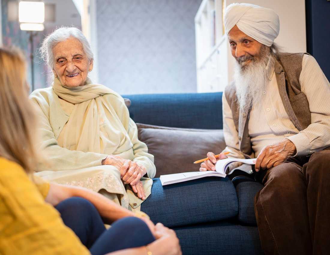 A group of three people in a care home are talking. They are an older man with a long white beard and turban, an older lady with grey hair wearing a cream top, and a younger lady who can only be seen from behind.