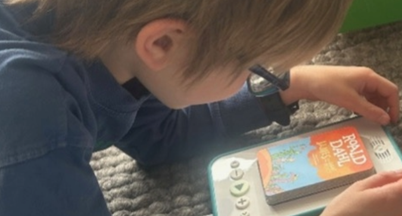 A young boy lies on a rug, focused on a handheld audio player in front of him as he carefully adjusts a book card. He wears glasses and appears absorbed in the activity, with the device positioned close to him while he listens and interacts with it.