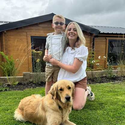 Riley, a young boy with severe sight loss and a beneficiary of BWBF, standing in the garden with his mum Jeanette, her arms wrapped around him. Buddy dog Bryce sits in front of them.