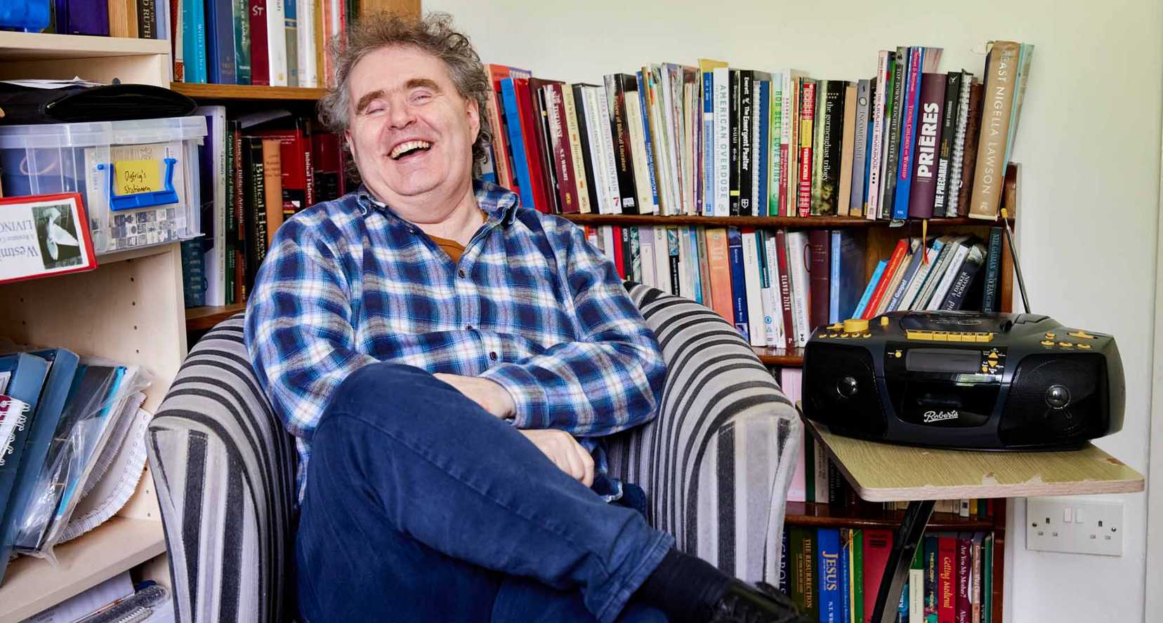 BWBF service user Dyfrig sits in a chair in a room with bookshelves. He is laughing and his Concerto 3 radio from BWBF is on a table next to him