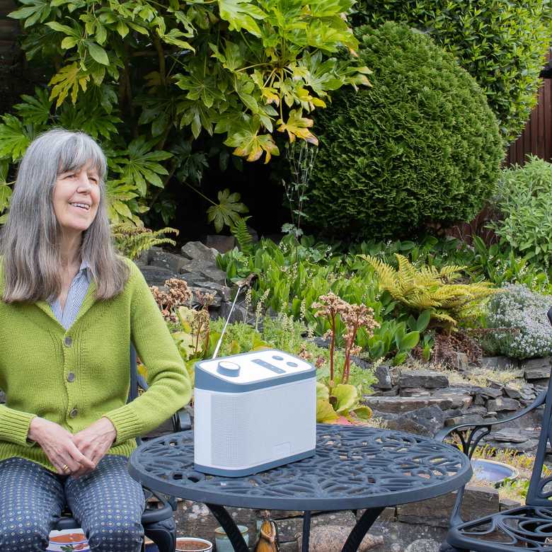 BWBF service user Janet sits at a garden table with the Relish radio we gave her. She has long grey hair and wears a green cardigan.