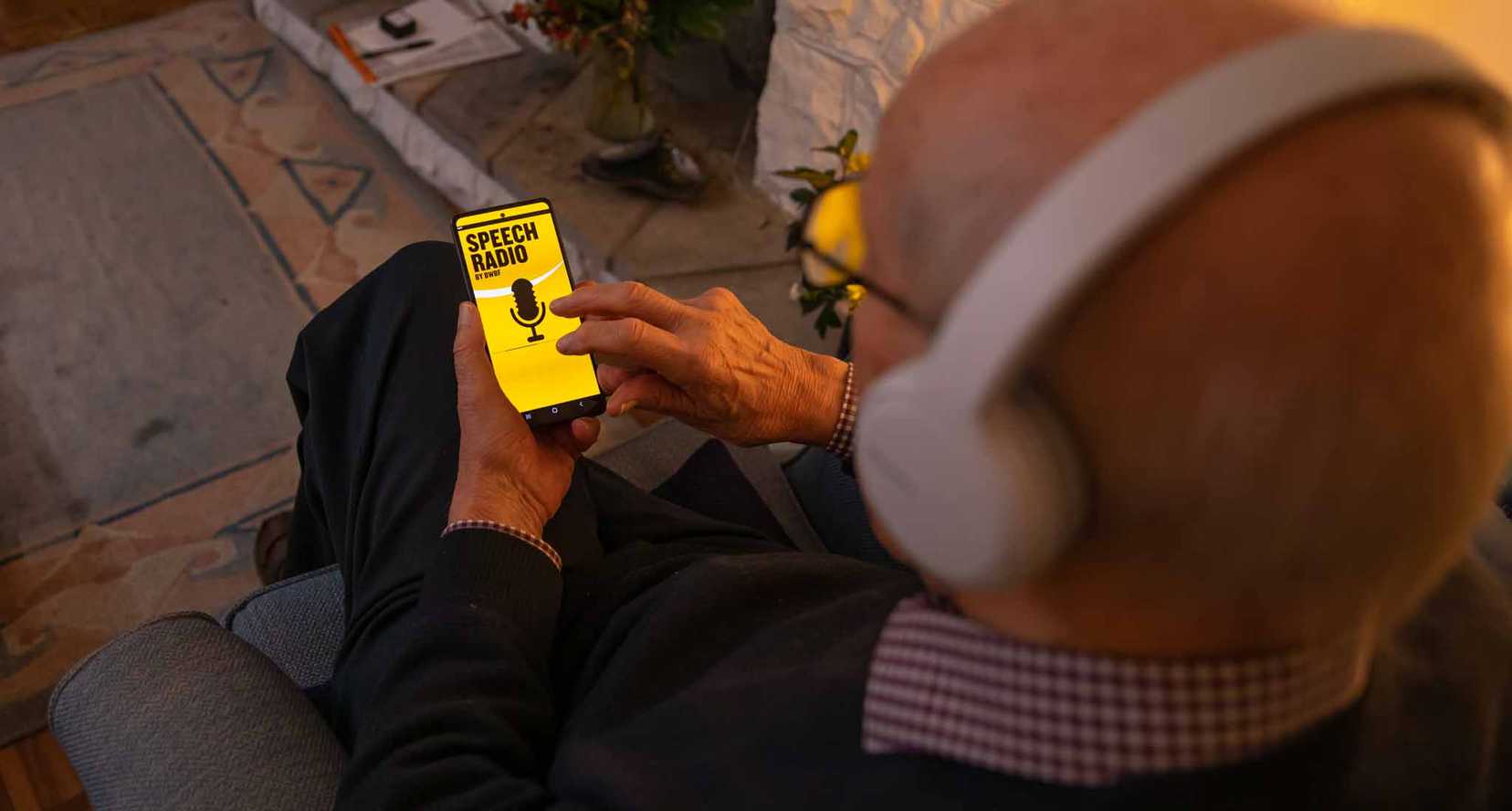 A man is browsing BWBF's Speech Radio app in a dimly lit room. He is wearing headphones.