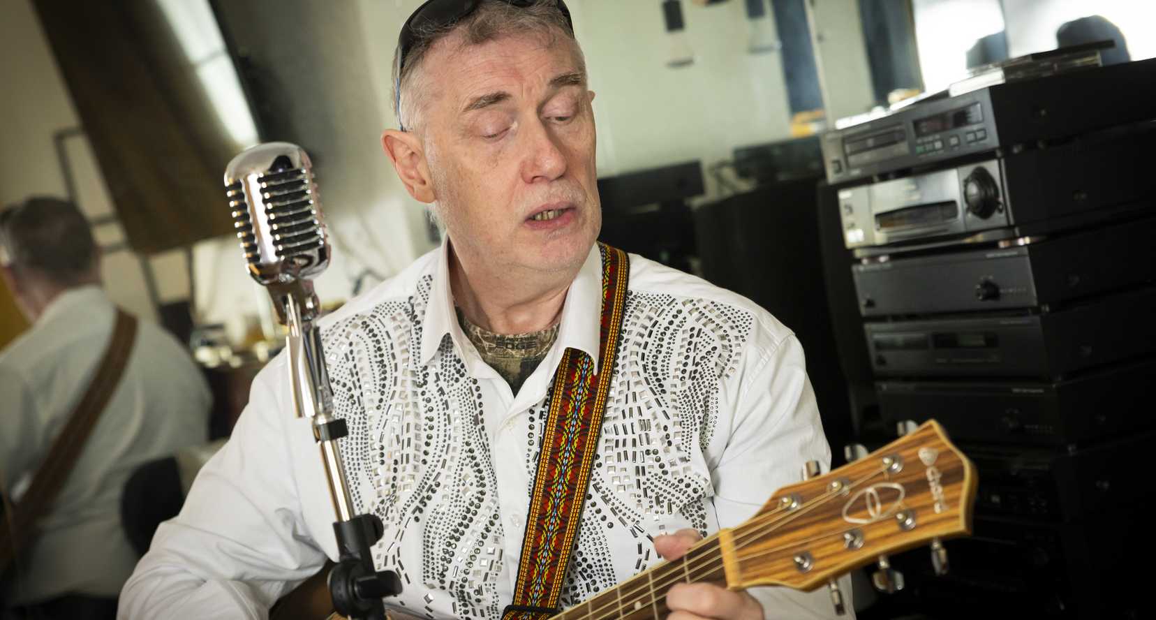 Paul Quigley, musician and performer. Paul lives with sight loss and is a beneficiary of BWBF. He is seated playing his guitar with a standalone microphone in front of him.