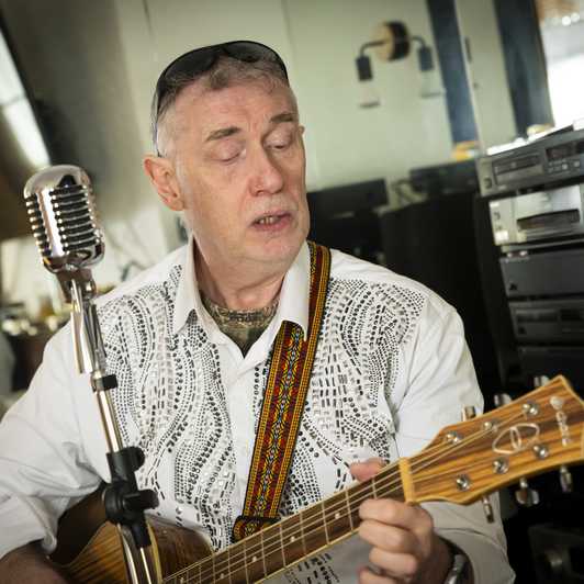 Paul Quigley, musician and performer. Paul lives with sight loss and is a beneficiary of BWBF. He is seated playing his guitar with a standalone microphone in front of him.