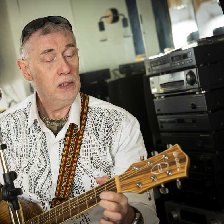 Paul Quigley, musician and performer. Paul lives with sight loss and is a beneficiary of BWBF. He is seated playing his guitar with a standalone microphone in front of him.