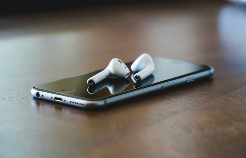 A mobile phone lies on black table surface. On top is a pair of cordless in-ear headphones.