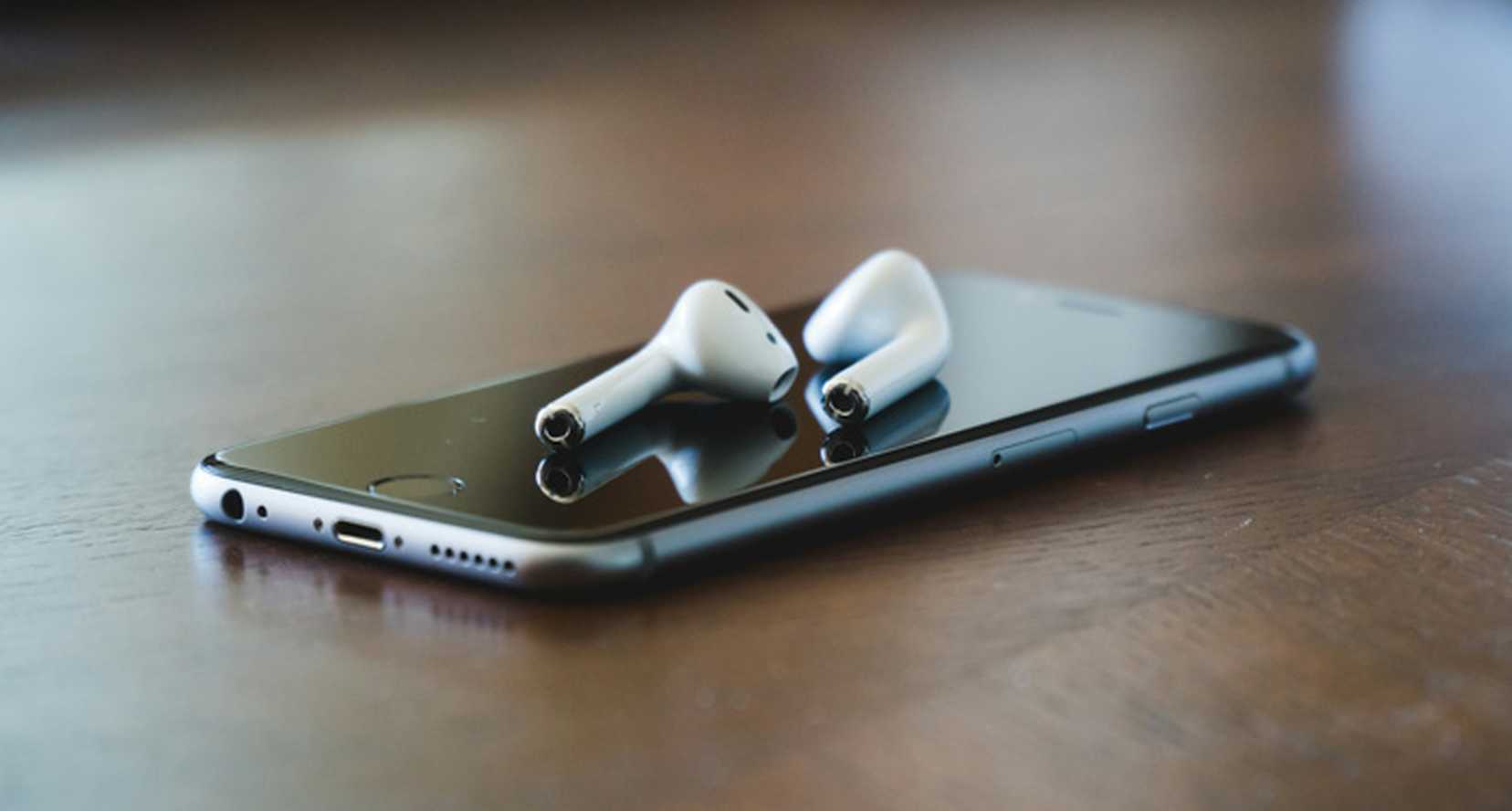 A mobile phone lies on black table surface. On top is a pair of cordless in-ear headphones.