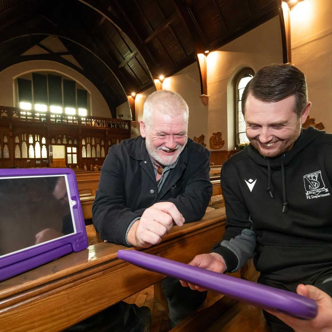 Two men hold iPads with purple cases and discuss. They are smiling.