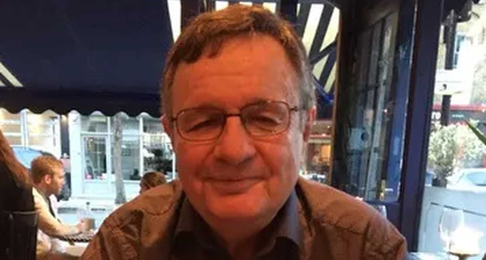 Paul Easton, who passed away in February 2023, is sitting smiling to camera. He wears glasses and has short dark hair.