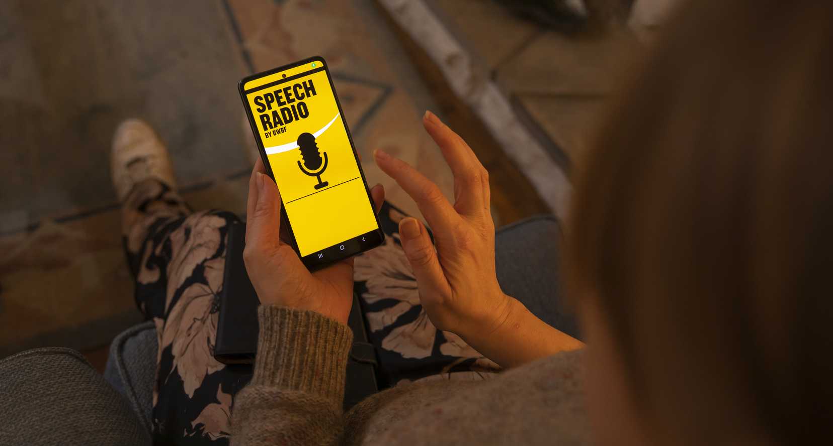 A person is browsing BWBF's Speech Radio app in a dimly lit room. The screen is lit up in yellow with the words 'Speech Radio' and an image of a microphone in black.