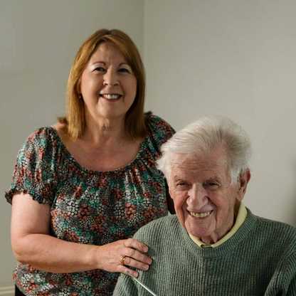 Thomas sits at a table smiling with his Relish radio from BWBF in front of him. Yvonne stands behind him also smiling, with her hand on his shoulder.