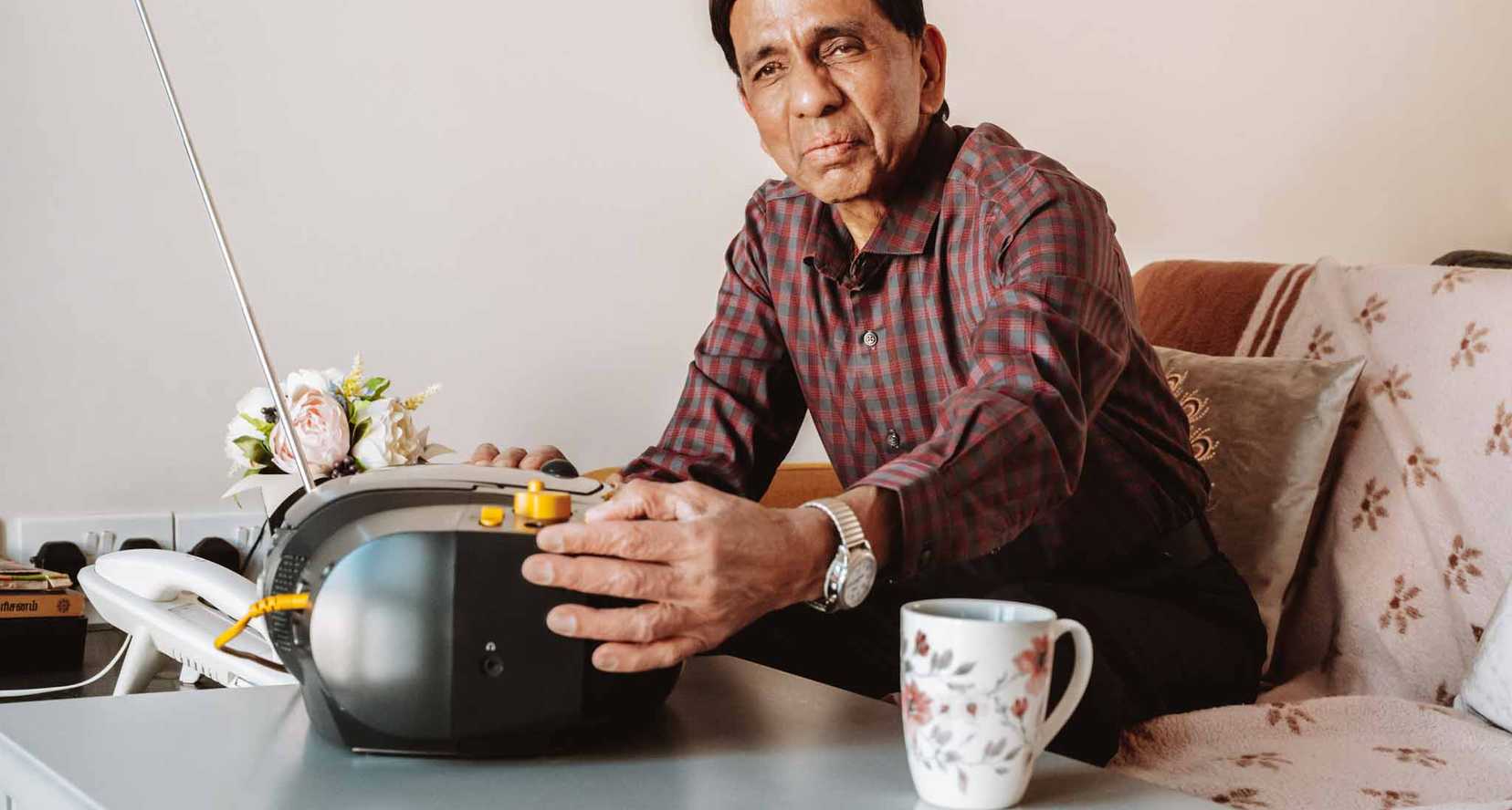 BWBF service user Krisha sits on a sofa. His hand is resting on his Concerto 3 radio from BWBF, which is on the table in front of him.