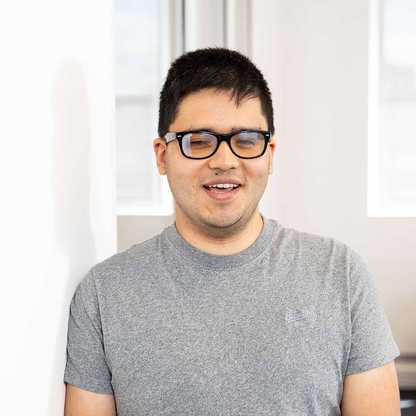 Suresh Kuwar, Web Developer for BWBF, is standing in an office space and smiling. He is wearing a grey t-shirt and glasses and has short, dark hair.
