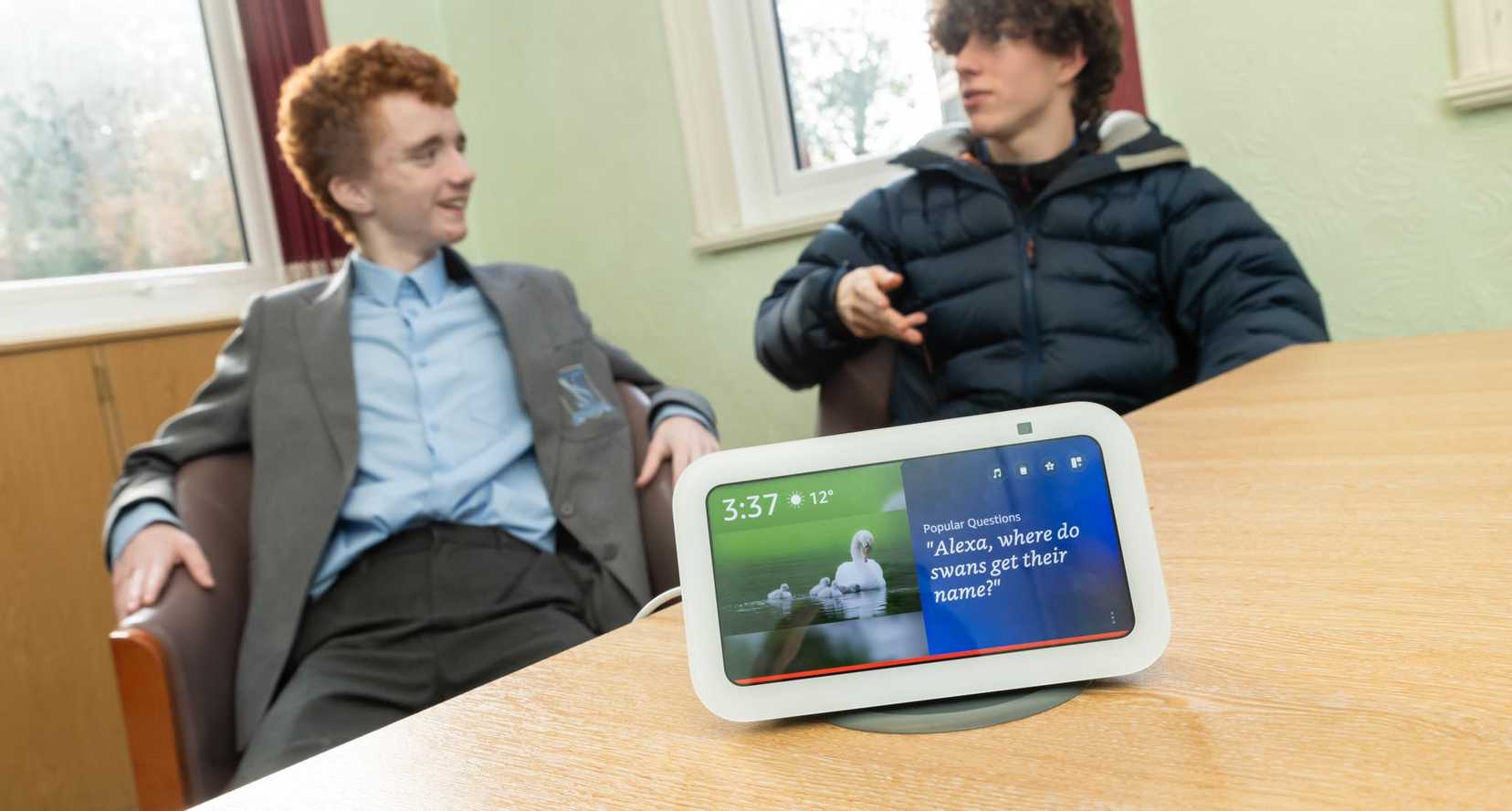A white Alexa Show device with two teenage boys in the background, discussing something.