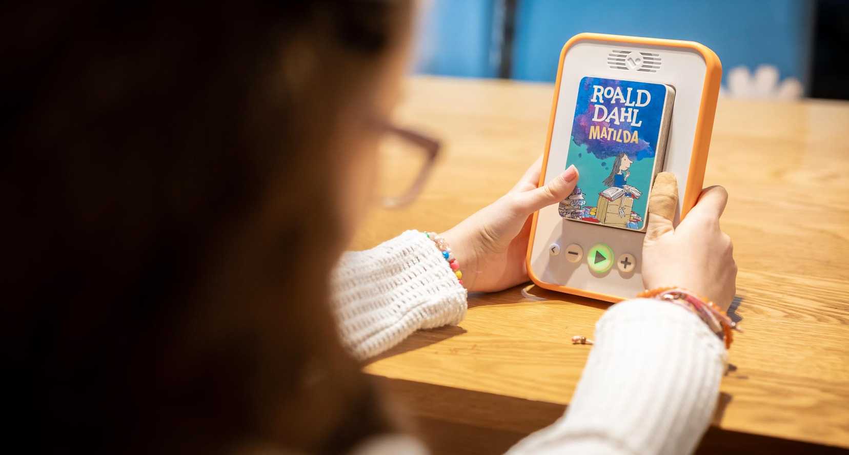 BWBF service user Annabel with her Voxblock audio book reader. Her hands are resting on the table and inside the slot in the reader is a copy of Roald Dahl's Matilda.