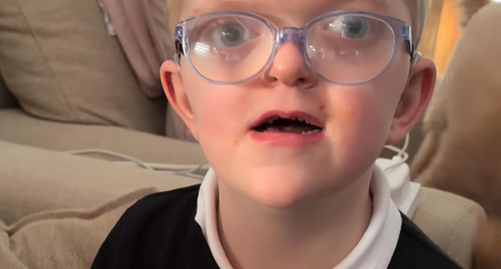 Close-up of Chip, 6, who has sight loss and complex needs. He has short blond hair, is wearing large, clear-framed glasses and a black top with a white collar. He is sitting indoors on a sofa and looking slightly upward.