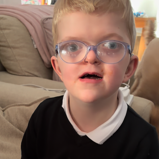 Close-up of Chip, 6, who has sight loss and complex needs. He has short blond hair, is wearing large, clear-framed glasses and a black top with a white collar. He is sitting indoors on a sofa and looking slightly upward.