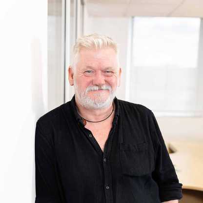 Steven Poole, Service Delivery Manager for BWBF, is leaning on a wall and smiling. He has wide grey hair and stubble is wearing a black shirt.