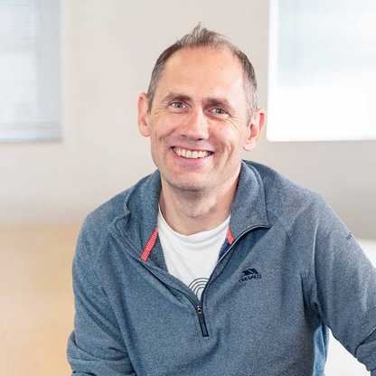 Simon Luget, Financial Controller for BWBF, sits on a table and is smiling. He has short grey hair and is wearing a bale blue casual top.
