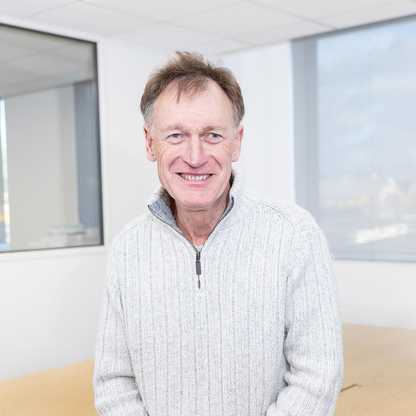Mark Noble, BWBF Trustee, photographed in 2024. He is pictured smiling, sitting on a desk in an office and he is wearing a cream woollen jumper and grey trousers.