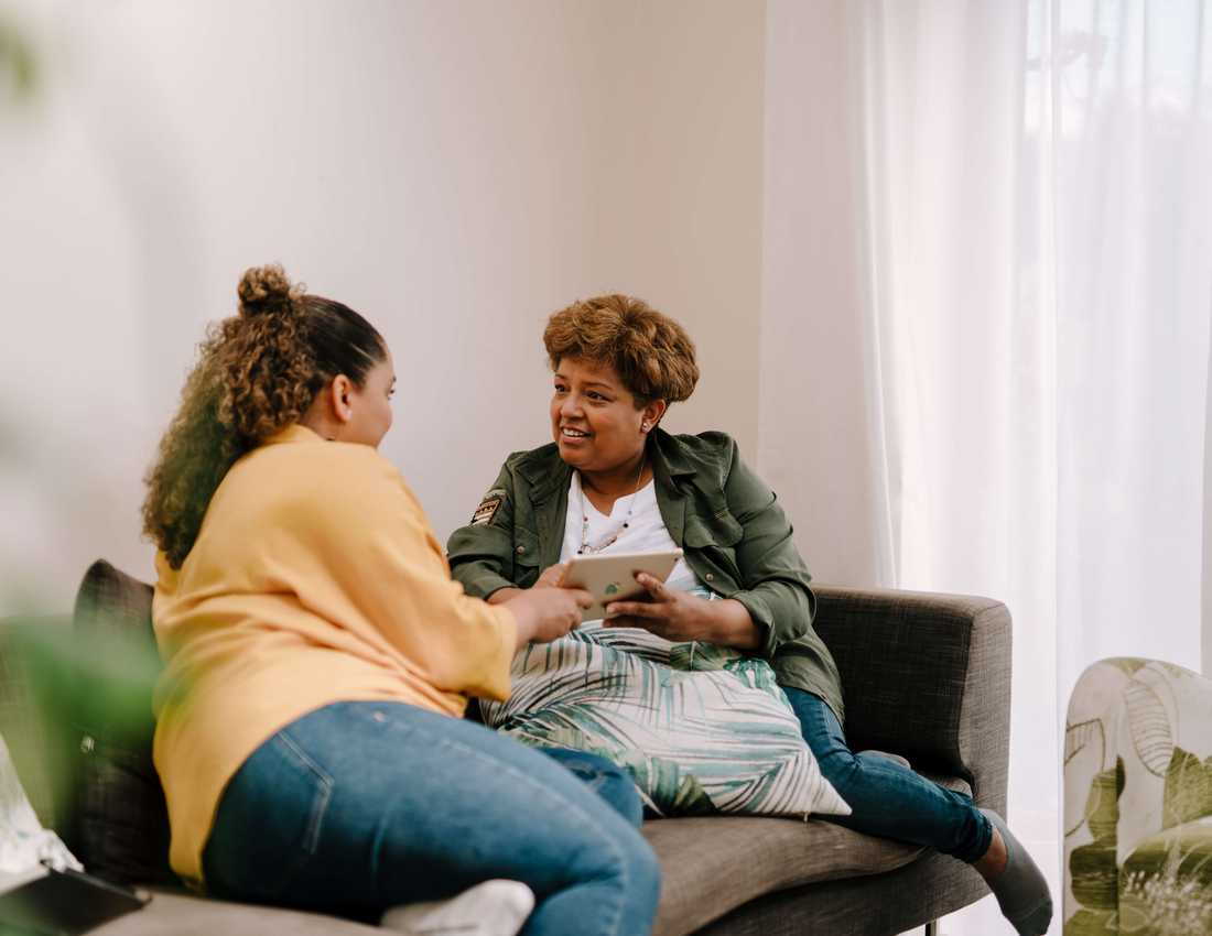 Two women sit on a sofa. They are smiling as they look at an iPad together.
