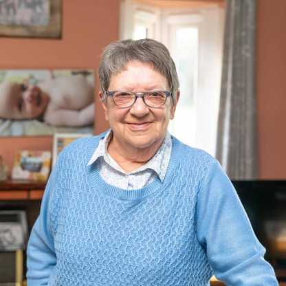 Norma stands in her living room smiling. She has short grey hair and glasses.