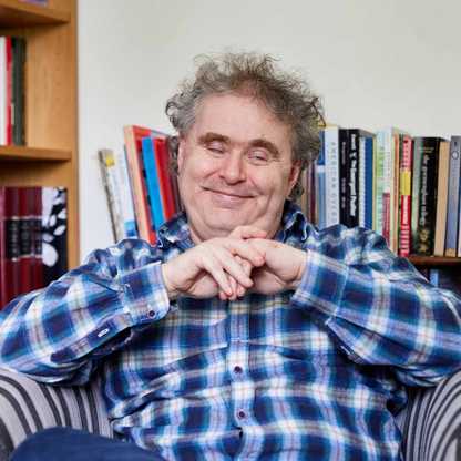 BWBF service user Dyfrig sits in a chair in a room with bookshelves. He is smiling and holds his hands together in front of him.