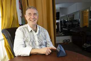 Paul Quigley, musician and performer. Paul lives with sight loss and is a beneficiary of BWBF. He is seated, smiling, as he engages with an Echo Show device on the table in front of him.