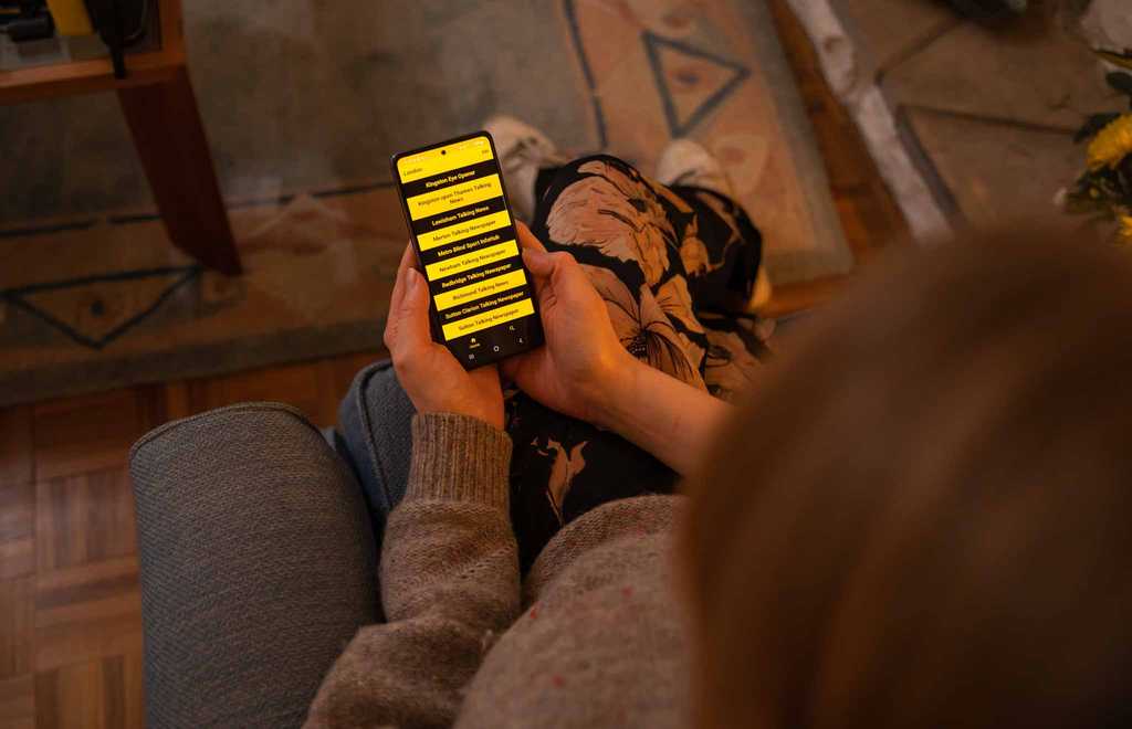 A BWBF service user holds a phone with BWBF's Speech Talking Newspaper app open on screen. A series of publications are listed in a yellow and black high-contrast design.