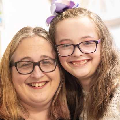 BWBF service user Annabel with her mum smiling to camera. Both wear glasses and Annabel has a bow in her hair.