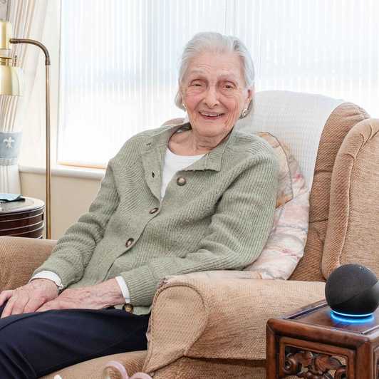Doreen, who got a free Echo Dot from BWBF at the age of 95, smiles as she sits in an armchair with her device next to her