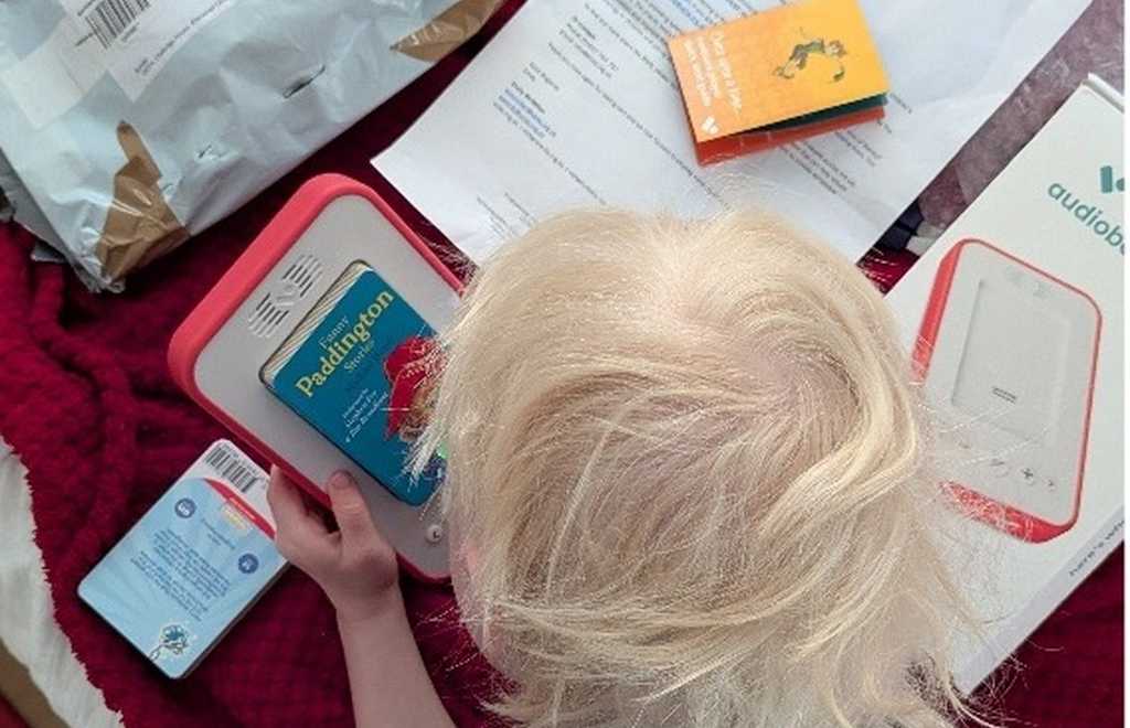 A young child with light blond hair sits on a red blanket, holding a Voxblock, an audiobook player with a “Paddington” book cover in it. Around him are opened packages, paperwork, and other audio equipment boxes, suggesting a recent delivery and setup,