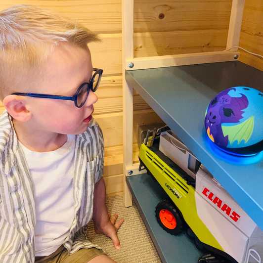 A boy, Riley, sits next to a shelf with his Echo Dot Kids smart speaker on it.