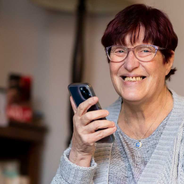 A middle aged woman smiles to camera as she holds her phone in her mobile phone up to her mouth.