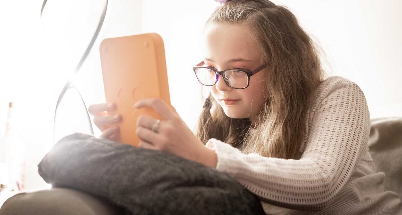 Annabel sits on her sofa reading her Voxblock audio book player. She wears glasses and has a bow in her hair.