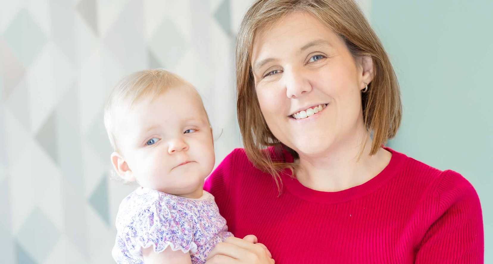 Young mother Laura smiles as she holds her baby daughter Daphne in her arms. She is wearing a vibrant red top.
