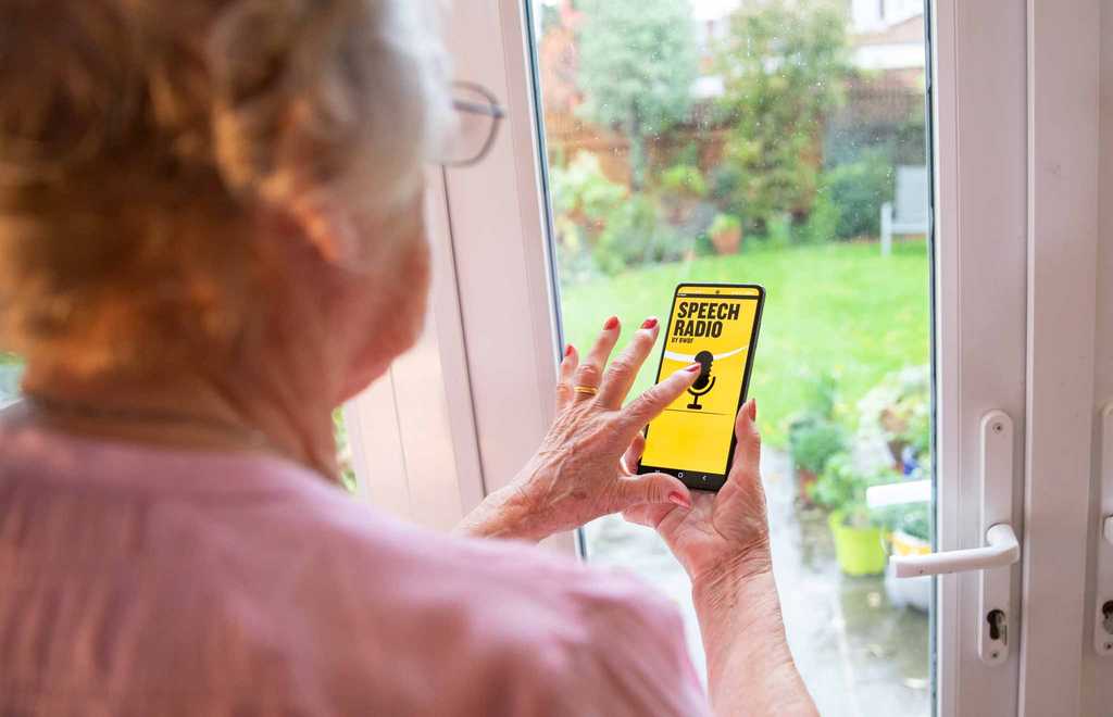 A woman stands in front of a window browsing BWBF's Speech Radio app. The app is black and yellow with an image of a microphone on it.