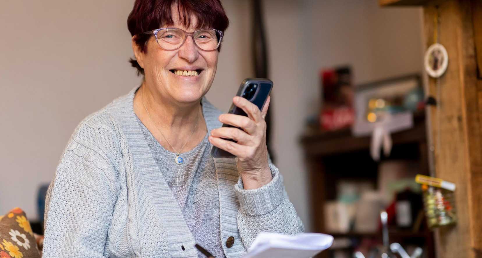 A woman stands in a kitchen smiling and as she talks on her phone which she is holding in front of her.