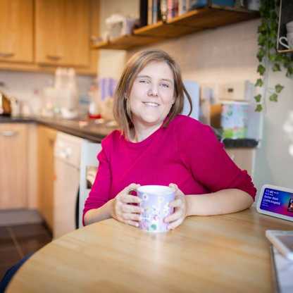 Laura at home in her kitchen smiling and with a cup of tea. On the table is her her free Alexa device.