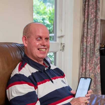 Brian, one of BWBF's app users, sits on a brown leather sofa smiling as he holds up his mobile phone.