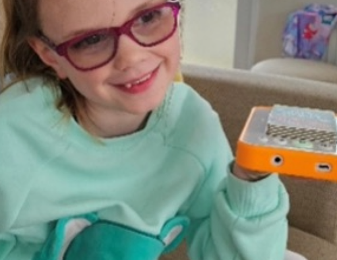 A young girl sits comfortably on a sofa, smiling as she holds a small audio player in her hand. Wearing glasses and casual clothes, she appears relaxed and engaged, with the device positioned as if she’s listening to or about to use it in a cosy home sett
