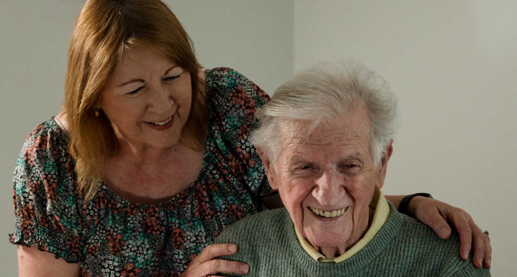 Thomas sits at a table smiling. His daughter Yvonne stands behind him looking at him and smiling, with her hands on his shoulder.