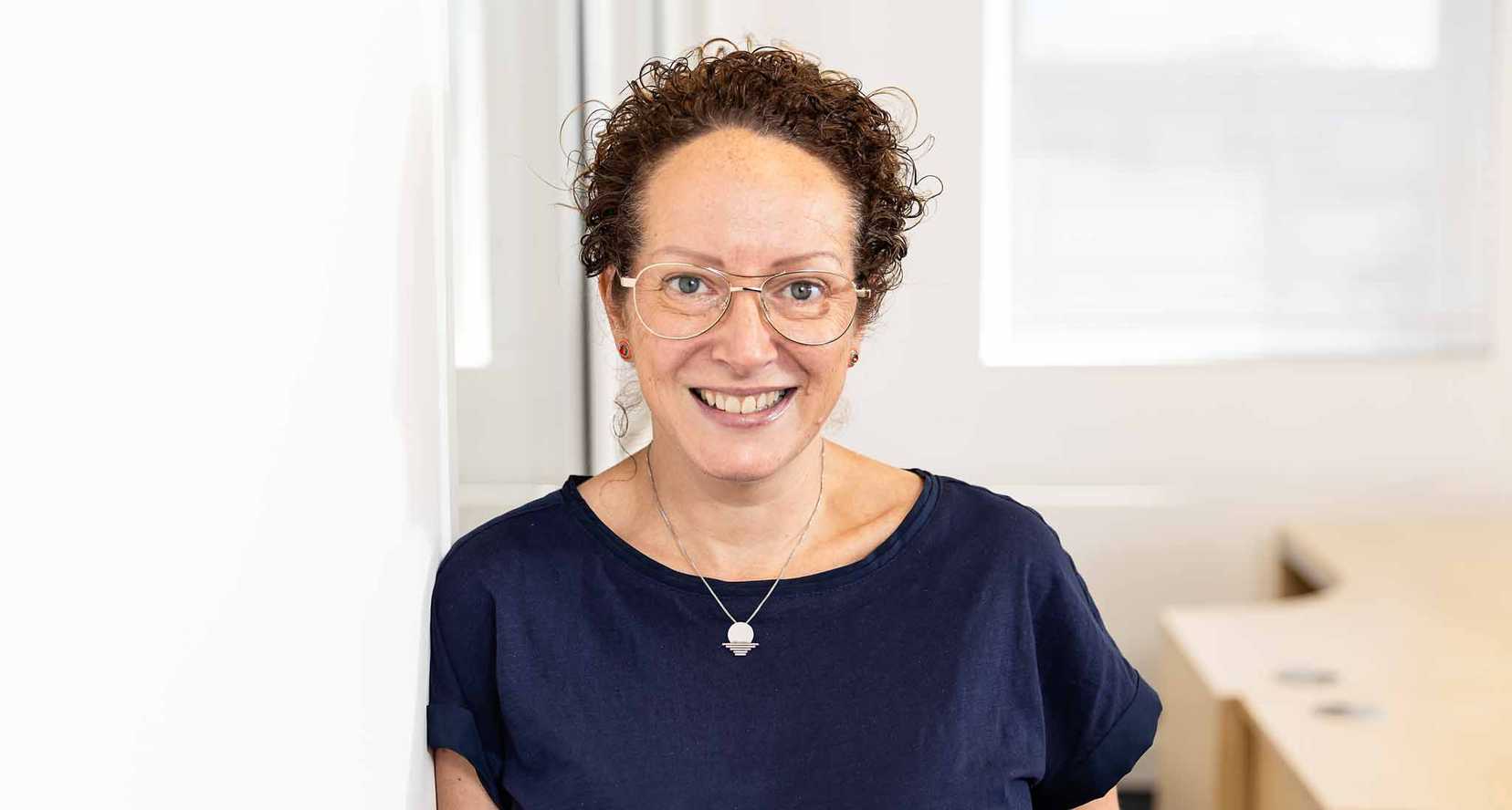 Julie Hocking, Individual Giving & Legacies Manager for BWBF, leans against a wall and is smiling. She wears glasses and her curly hair is tied up.