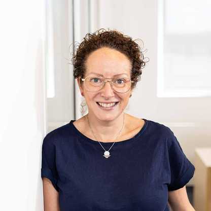Julie Hocking, Individual Giving & Legacies Manager for BWBF, leans against a wall and is smiling. She wears glasses and her curly hair is tied up.