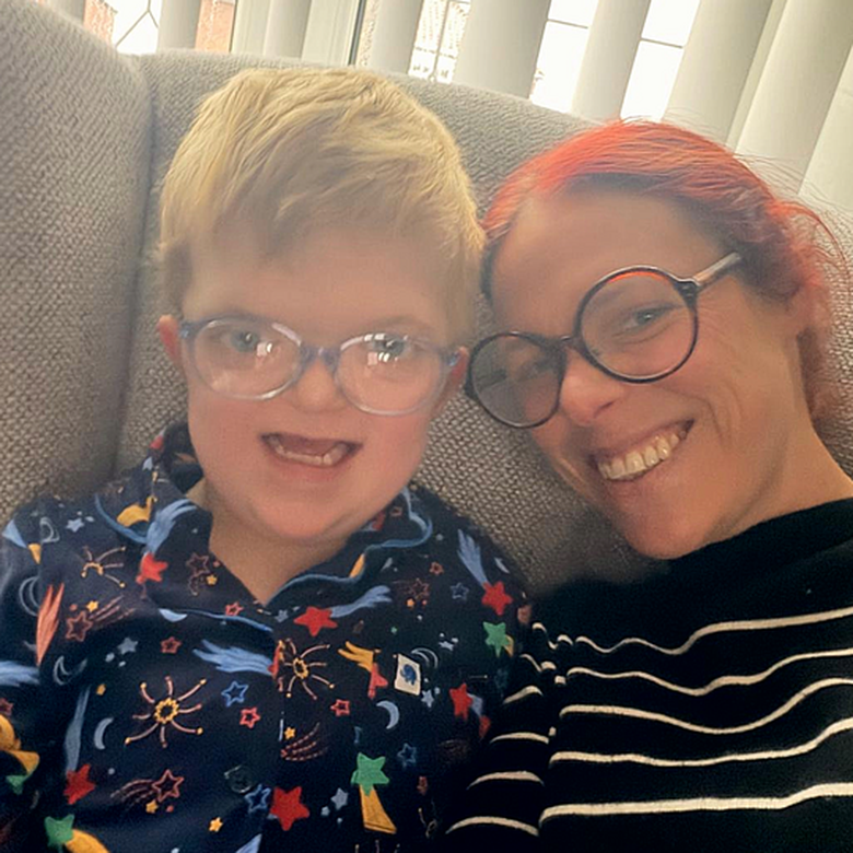 Close-up of Chip, 6, who has sight loss and complex needs, sitting on a sofa beside his mum. He has short blond hair and wears large, clear-framed glasses and patterned pyjamas, while his mum, with red hair and round glasses, leans in smiling.