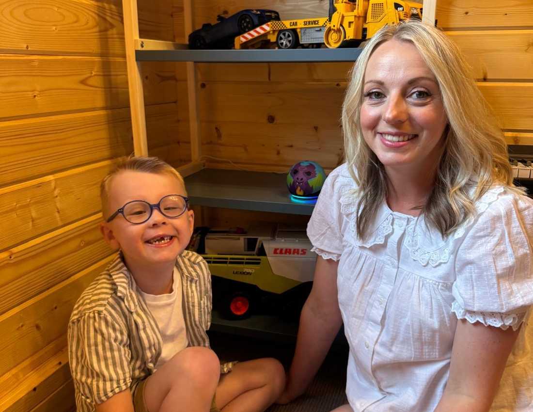 Riley and his mum are sitting on the floor smiling to camera. Behind them on the shelf is Riley's Echo Dot Kids smart speaker from BWBF.