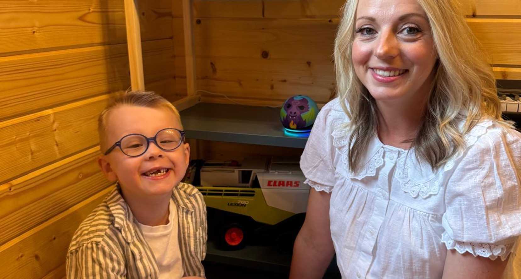 Riley and his mum are sitting on the floor smiling to camera. Behind them on the shelf is Riley's Echo Dot Kids smart speaker from BWBF.